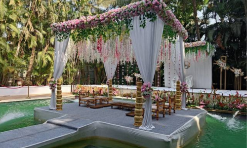 Floating wedding mandap on a water pool with white drapes and pink flowers, surrounded by lush greenery.